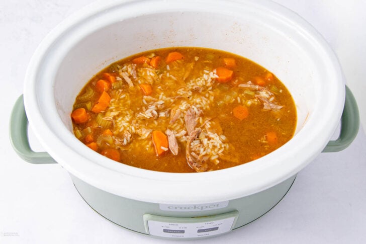 A slow cooker filled with soup containing shredded meat, rice, sliced carrots, and broth, sitting on a white surface.