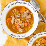 A bowl of soup with rice, carrots, celery, and meat on a white plate with the words “Let everything that has breath praise the Lord!” printed on the rim, next to two spoons on a yellow cloth.