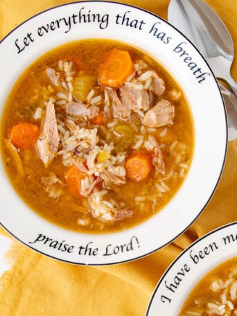 A bowl of soup with rice, carrots, celery, and meat on a white plate with the words “Let everything that has breath praise the Lord!” printed on the rim, next to two spoons on a yellow cloth.
