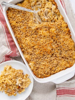 A white baking dish filled with creamy chicken casserole topped with golden breadcrumbs. A serving is on a small white plate nearby, with two forks resting in the dish on a beige and red-striped cloth.