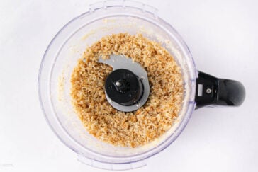 A food processor bowl filled with finely ground, light brown mixture, possibly breadcrumbs or crushed nuts, viewed from above on a white background.