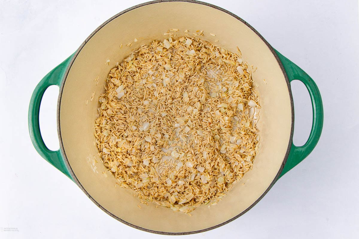 Top view of a green Dutch oven containing uncooked rice and chopped onions on a white surface.