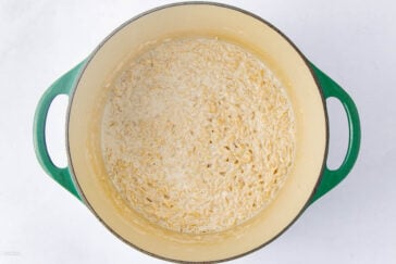 A green Dutch oven filled with creamy, cooked orzo pasta sits on a white background. The orzo appears soft and coated in a light sauce, with no additional ingredients visible.