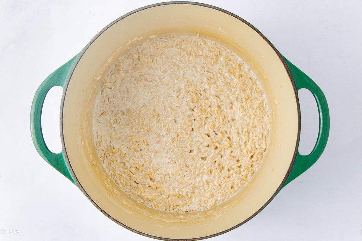 A green Dutch oven filled with creamy, cooked orzo pasta sits on a white background. The orzo appears soft and coated in a light sauce, with no additional ingredients visible.