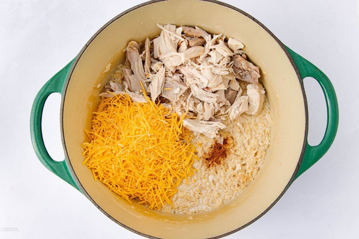 A green Dutch oven containing shredded chicken, grated cheddar cheese, cooked rice, and a sprinkle of seasoning, all arranged separately before mixing.