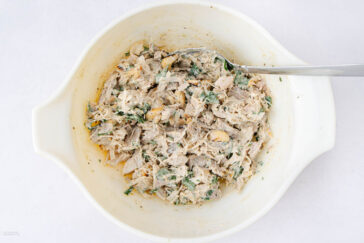 A white mixing bowl filled with a creamy chicken salad mixture, containing shredded chicken, herbs, possibly nuts, and a spoon resting inside the bowl.