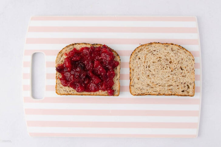 Two slices of whole grain bread on a pink and white striped cutting board; one slice has red jam spread on top, the other is plain.