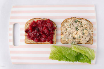 Two slices of bread on a striped cutting board—one topped with cranberry sauce, the other with creamy chicken salad. A piece of romaine lettuce lies in front, ready to be added.