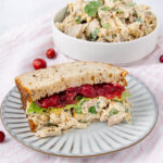 A sandwich on multigrain bread filled with chicken salad, lettuce, and cranberry sauce sits on a striped plate. In the background, a bowl of chicken salad is visible. Both are set on a light pink striped cloth.