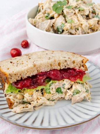 A sandwich on multigrain bread filled with chicken salad, lettuce, and cranberry sauce sits on a striped plate. In the background, a bowl of chicken salad is visible. Both are set on a light pink striped cloth.