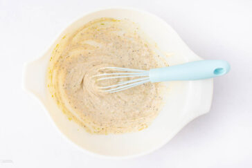 A white mixing bowl with a blue-handled whisk inside, containing a light batter with visible specks, placed on a white surface.