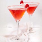 Two martini glasses filled with a pink cocktail, each glass rimmed with red sugar and garnished with a raspberry. The glasses are placed on a white surface with a soft white cloth underneath.