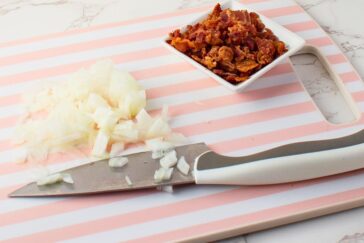A knife with a white handle rests on a pink and white striped cutting board with chopped onions beside it. A small square dish of chopped cooked bacon sits nearby.