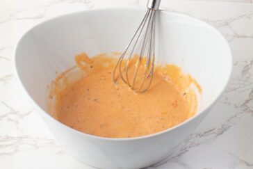 A metal whisk rests in a white bowl filled with a creamy, orange-colored sauce or dressing, set on a white marble surface.