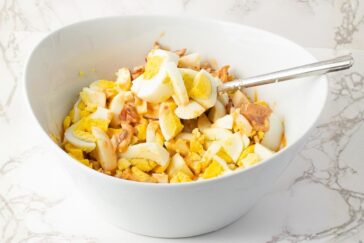 A white bowl filled with chopped hard-boiled eggs mixed with what appears to be pieces of tuna, with a fork resting inside. The bowl is on a white marble surface.