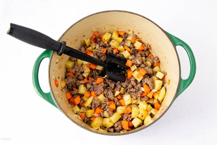 A green Dutch oven filled with diced potatoes, carrots, ground beef, and seasonings sits on a white surface. A black spatula rests inside the pot, mixing the ingredients.