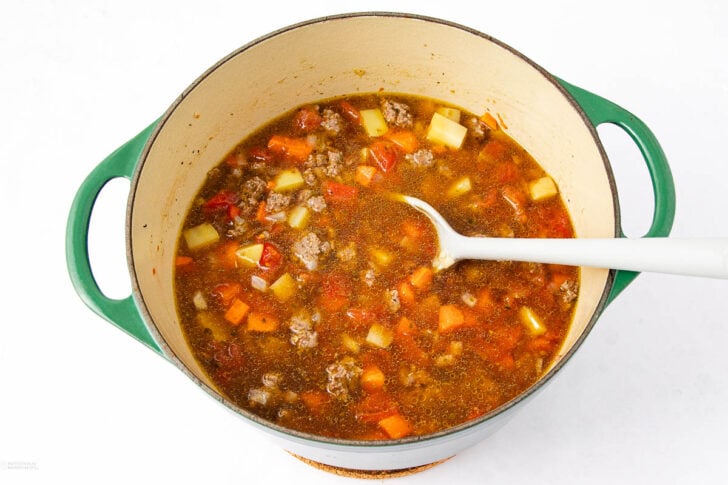 A green pot filled with beef and vegetable soup, including chunks of carrots, potatoes, and ground beef, with a white spoon resting inside.