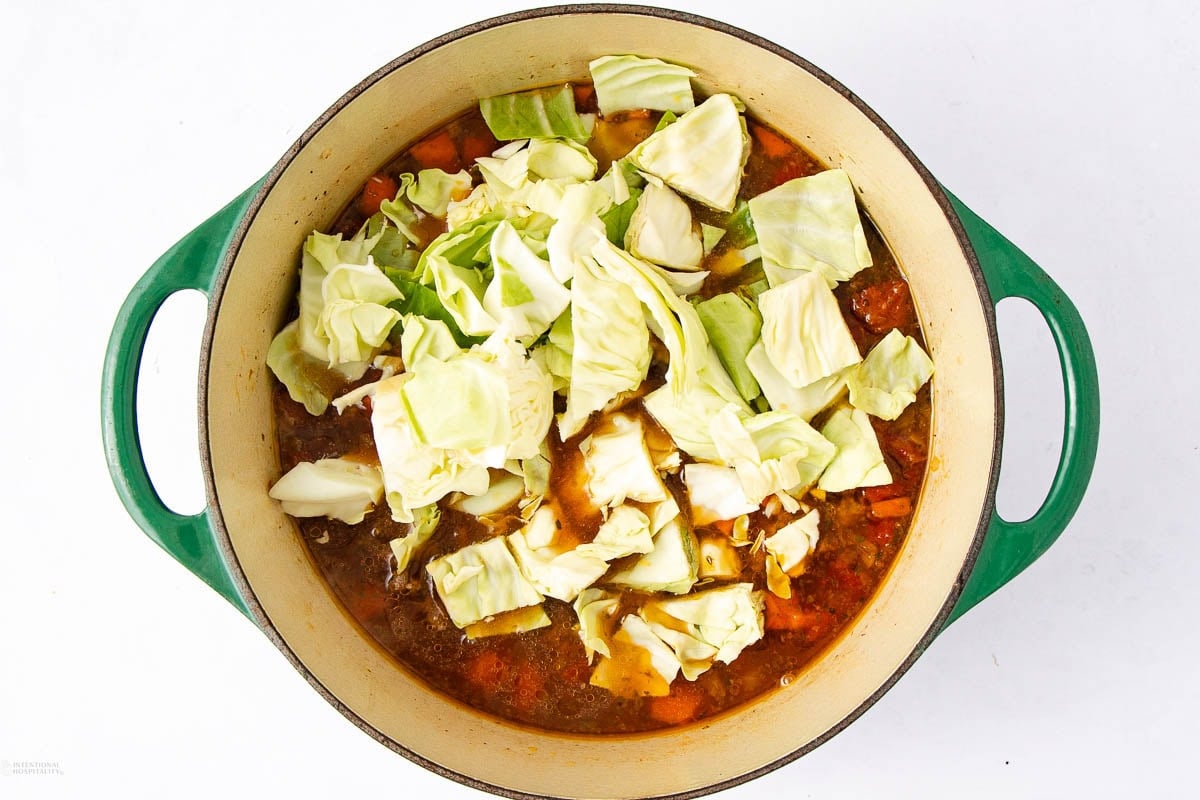 A green Dutch oven filled with soup, topped with large chunks of raw cabbage, sits on a white background. The soup base beneath the cabbage appears to contain tomatoes and possibly other vegetables.