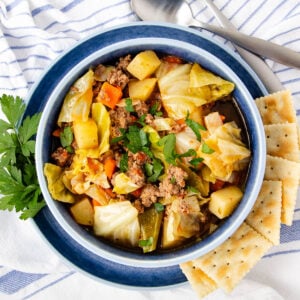 A bowl of cabbage soup with ground beef, potatoes, carrots, and parsley, served with saltine crackers on the side, and a sprig of parsley for garnish. Spoons and a striped towel are in the background.