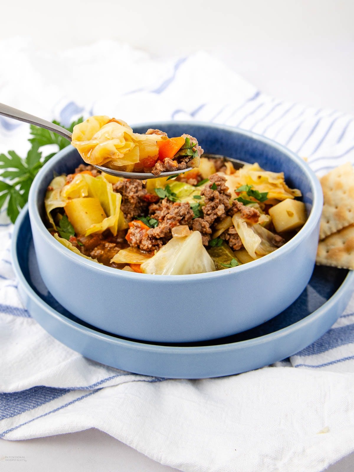 A blue bowl filled with cabbage soup containing ground beef, potatoes, and vegetables. A spoonful of soup is held above the bowl, with crackers on the side and a sprig of parsley nearby.
