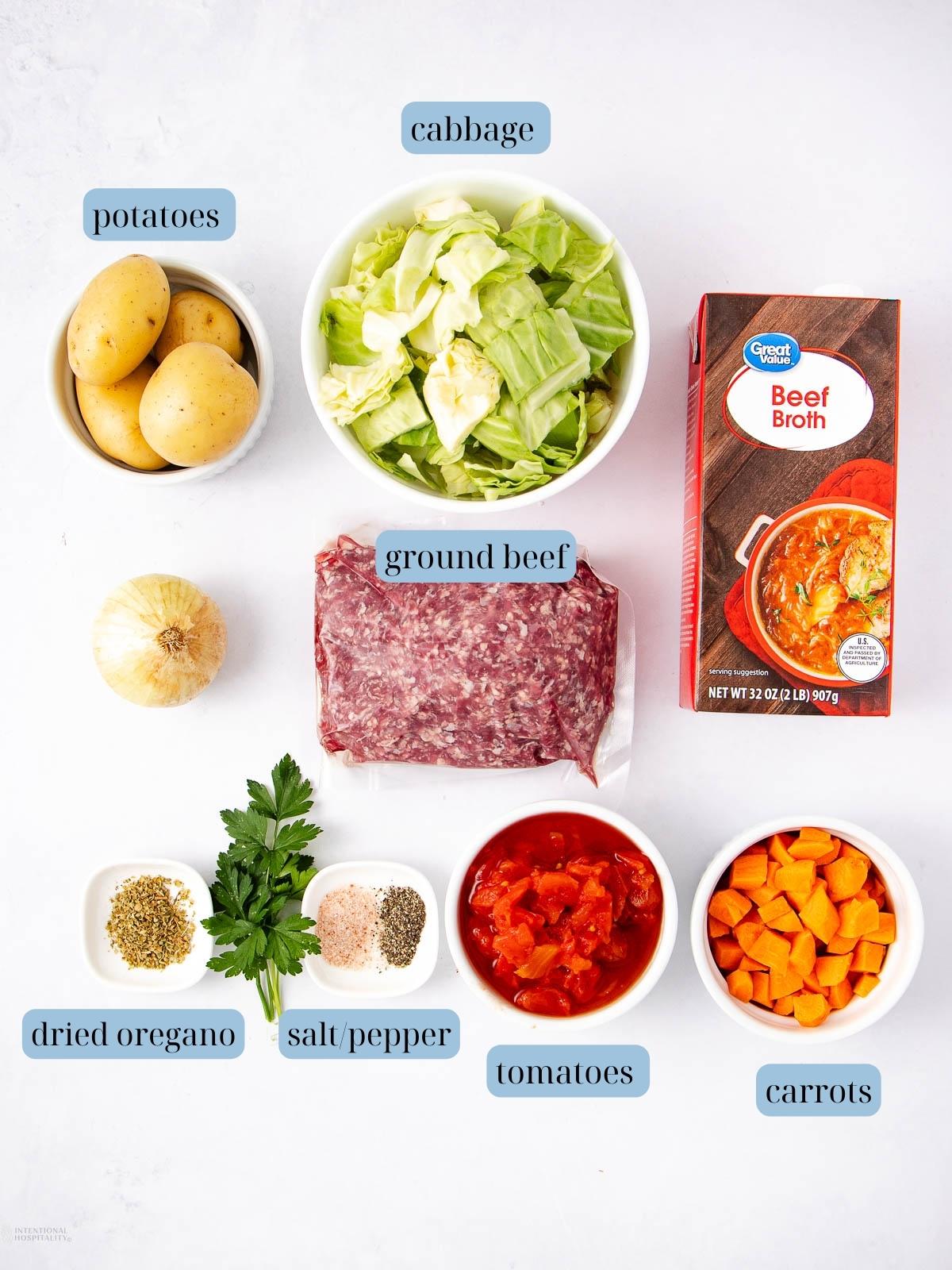 A top-down view of ingredients for soup: a bowl of potato pieces, chopped cabbage, a carton of beef broth, whole onion, ground beef, bowls of spices, fresh parsley, diced tomatoes, and diced carrots, all labeled.