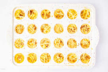 A white, speckled muffin tray filled with mini unbaked egg bites, each topped with shredded cheese and bits of vegetables or meat, is viewed from above against a white background.