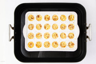 A baking tray with 24 mini quiches, each topped with shredded cheese and seasoning, sits inside a larger black roasting pan with metal handles on both sides.