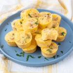 A blue plate holds a stack of mini egg muffins, garnished with chopped herbs, set on a yellow and white checkered cloth.