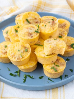A blue plate holds a stack of mini egg muffins, garnished with chopped herbs, set on a yellow and white checkered cloth.