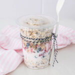 A clear plastic cup filled with yogurt, fresh berries, and granola, topped with a lid. A white spoon is attached to the cup with black-and-white twine. A pink and white striped cloth is in the background.