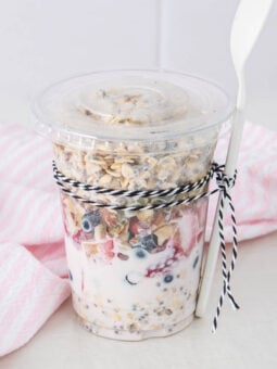A clear plastic cup filled with yogurt, fresh berries, and granola, topped with a lid. A white spoon is attached to the cup with black-and-white twine. A pink and white striped cloth is in the background.