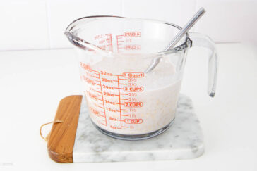 A glass measuring cup filled with a milky liquid and oats, with a metal spoon inside. The cup sits on a marble and wood cutting board against a white background.