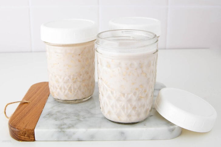 Three glass jars filled with creamy overnight oats sit on a marble and wood cutting board. Two jars have white lids on, while one jar has the lid off and placed nearby. The background is a white tiled wall.