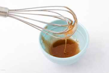 A metal whisk with brown liquid drips above a light blue bowl containing the same brown mixture, all set against a white background.