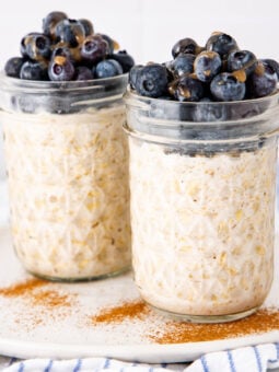 Two glass jars filled with overnight oats, topped with fresh blueberries and a sprinkle of cinnamon, sit on a white plate with a striped cloth in the background.