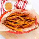 A red basket lined with parchment paper holds crispy sweet potato fries. Next to it is a small white bowl filled with seasoning, placed on a red-and-white striped towel.