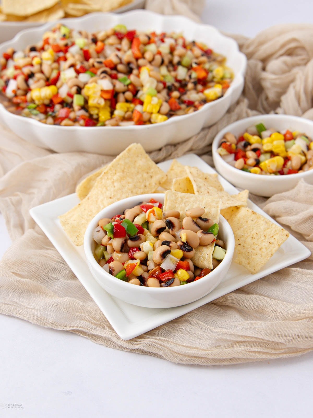 A square white plate holds a small bowl of black-eyed pea salad with chopped vegetables and three tortilla chips. Behind it are two more bowls with the same colorful salad, all set on a light beige cloth.