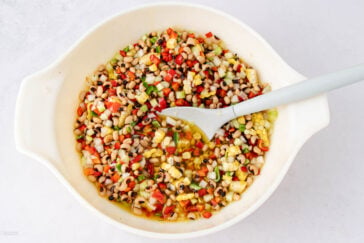 A white bowl filled with a colorful bean salad containing black-eyed peas, corn, diced red peppers, cucumbers, and onions, with a white serving spoon resting inside.