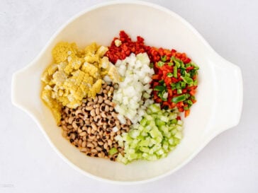 A white bowl containing chopped corn, red bell pepper, green bell pepper, onion, celery, and black-eyed peas arranged in separate sections on a white surface.