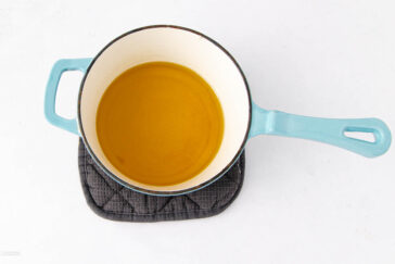 A blue-handled pot filled with oil sits on a black pot holder against a white background.