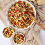 A large white bowl and two small bowls hold colorful Texas caviar made of black-eyed peas, corn, peppers, and onions. A wooden spoon rests in the main bowl. Beige fabric and tortilla chips surround the dishes.