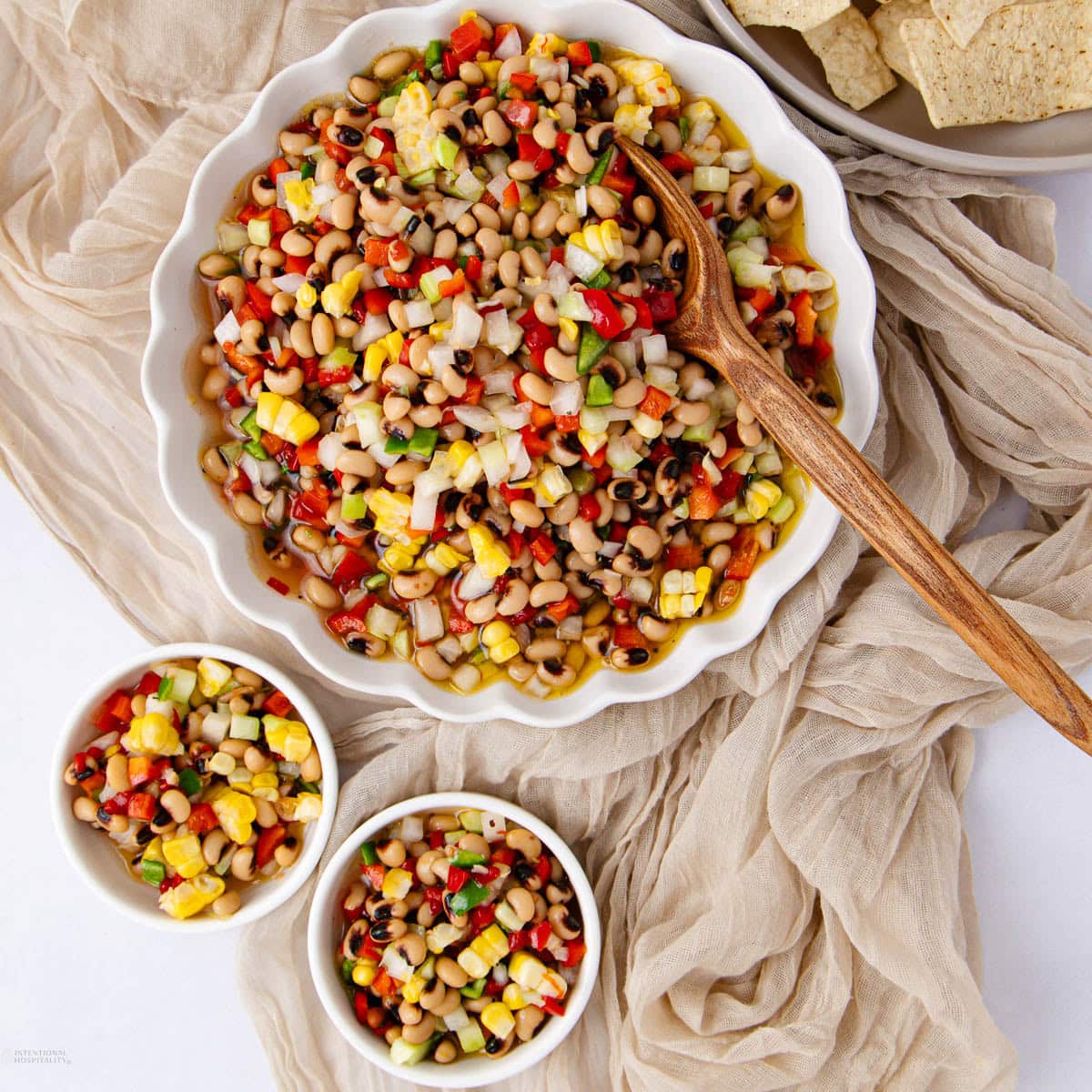 A large white bowl and two small bowls hold colorful Texas caviar made of black-eyed peas, corn, peppers, and onions. A wooden spoon rests in the main bowl. Beige fabric and tortilla chips surround the dishes.