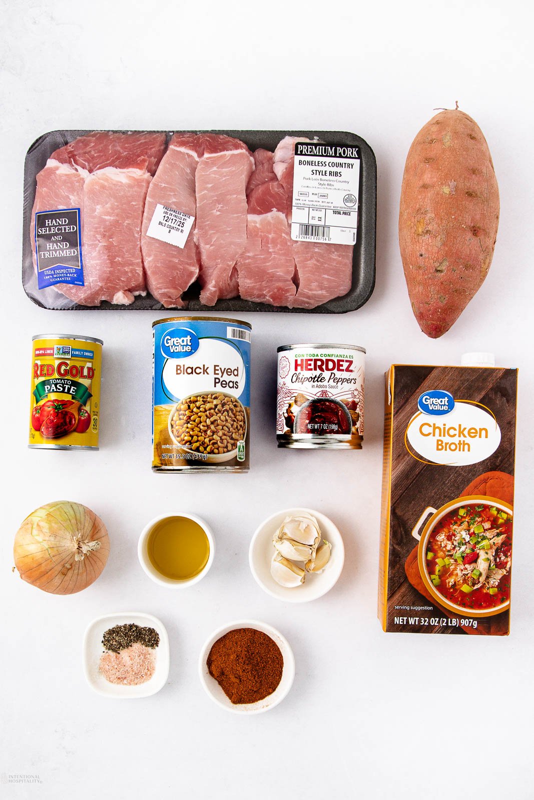 Raw pork chops, a sweet potato, a can of tomato paste, canned black-eyed peas, canned chipotle peppers, a carton of chicken broth, a yellow onion, olive oil, garlic cloves, and bowls of spices on a white background.