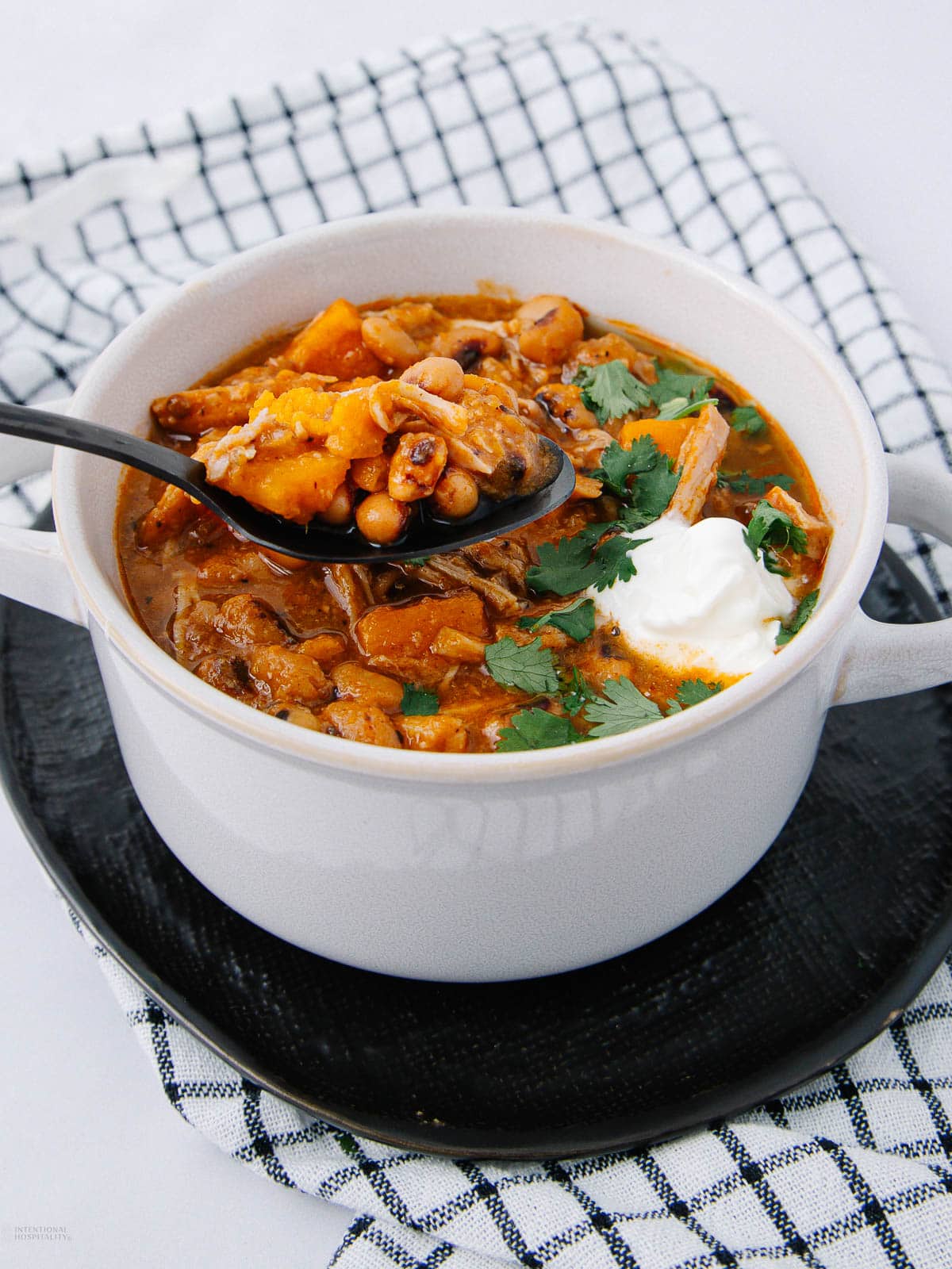 A white bowl of stew with beans, chunks of meat, and vegetables, topped with fresh cilantro and a dollop of sour cream, sits on a black plate with a checkered cloth underneath. A spoon is lifting a portion of the stew.