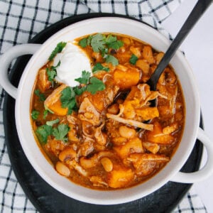 A white bowl filled with hearty stew featuring beans, chunks of orange squash, shredded meat, a dollop of sour cream, and fresh cilantro, set on a black tray over a checkered cloth.