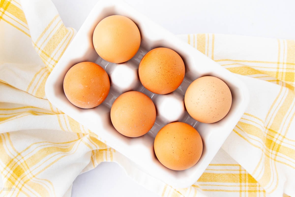 A white ceramic egg holder with six brown eggs sits on a white surface, next to a yellow and white checkered kitchen towel.