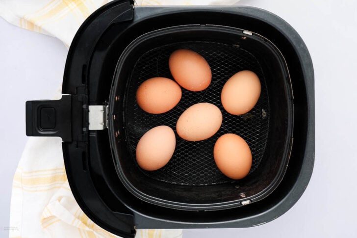 Six brown eggs are arranged inside the basket of a black air fryer, with a white and yellow striped towel partially visible beside it.