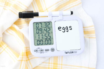 A digital kitchen timer with three time displays and a small whiteboard labeled "eggs" sits on a yellow and white checkered cloth. A black dry-erase marker rests on top of the timer.