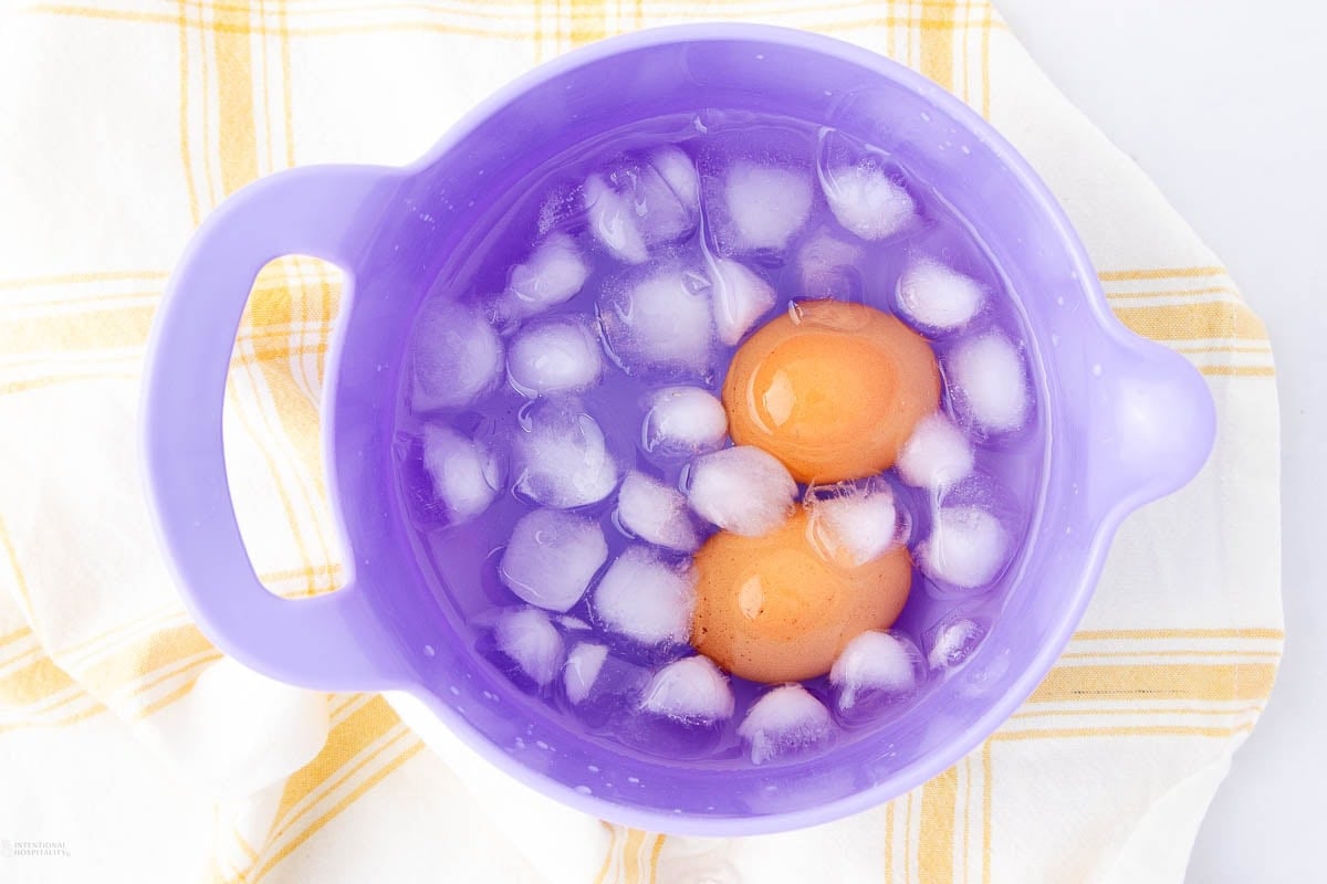 Two brown eggs in a purple bowl filled with ice cubes and water, resting on a white towel with yellow stripes.