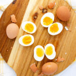 A wooden cutting board with three whole brown eggs, egg shells, and three halved boiled eggs showing different yolk textures, from soft and runny to firm. A yellow-checkered cloth is partially visible underneath.